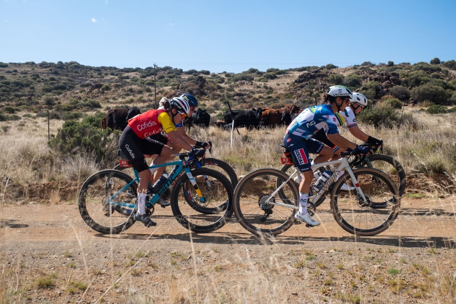 Ciclista con casco levantando la rueda delantera en un salto sobre una pista de grava seca, con un grupo de corredores con maillot naranja al fondo y montañas desérticas